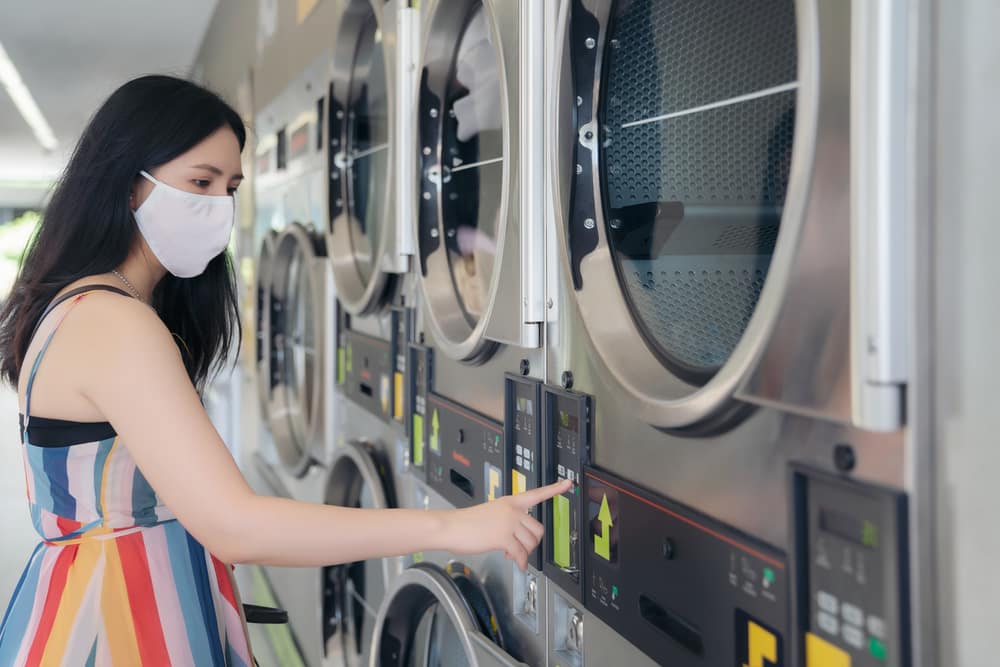 Young woman wearing mask using dryer at laundromat