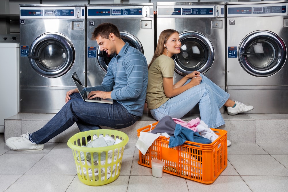 Young couple sitting in laundromat using laptop and earbuds