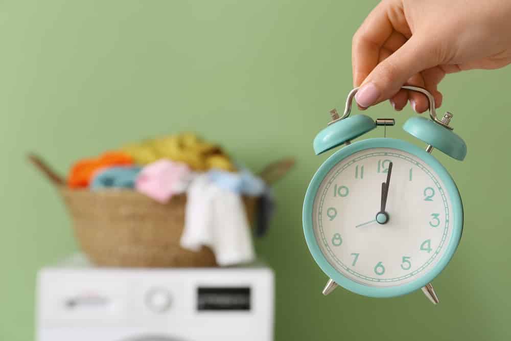 Hand holding up alarm clock in front of full laundry basket on washing machine